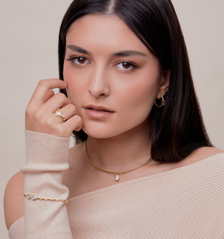 Woman wearing gold jewelry including a necklace, ring, and bracelet against a neutral background